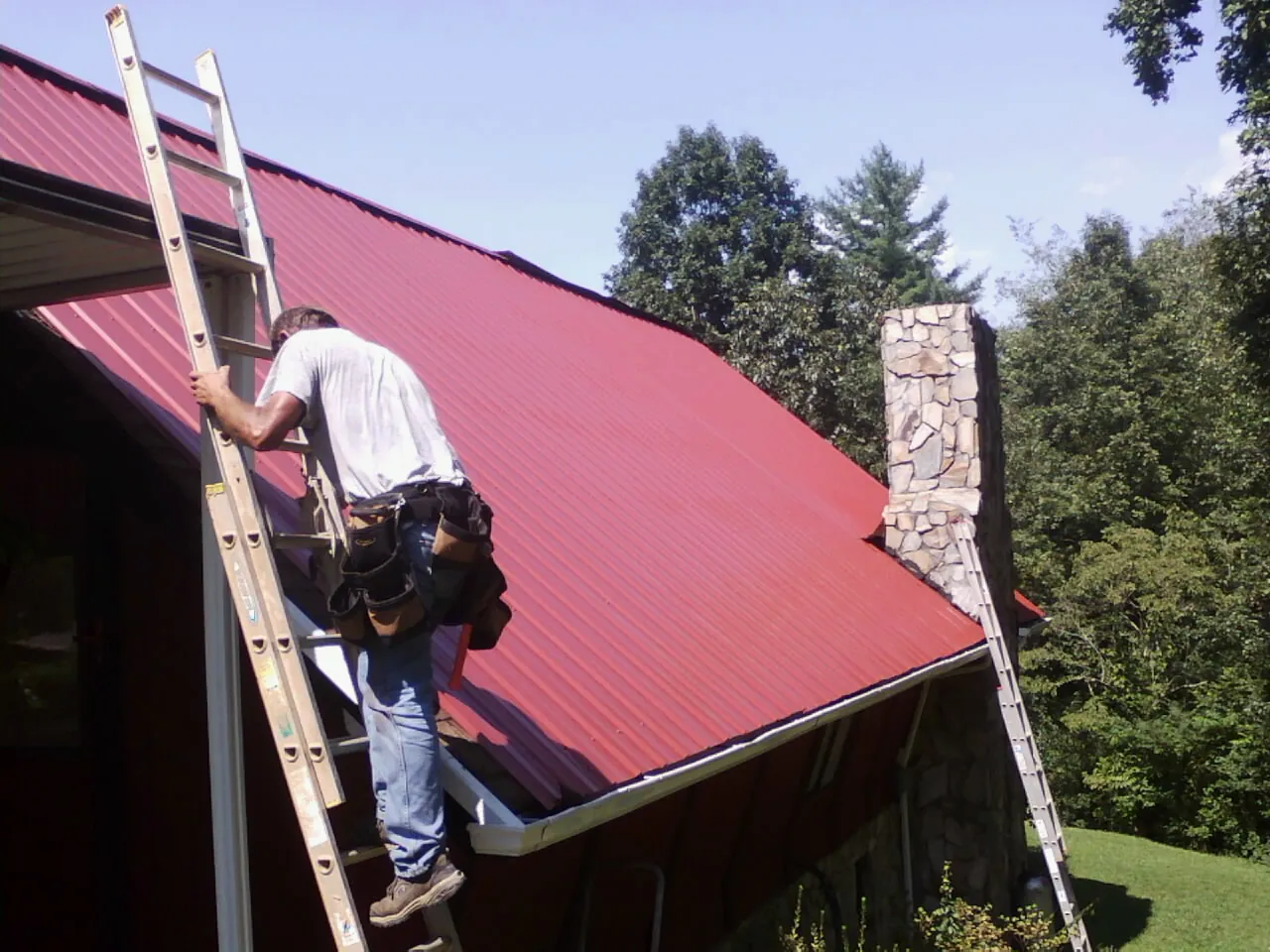 Roofer on a ladder beside a red metal roof and stone chimney — Soffit & Fascia Repair by Roofing Brothers in Bolton